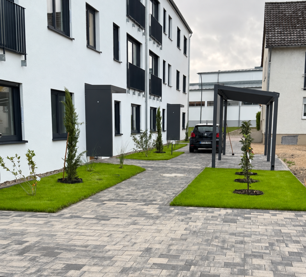 Außenanlage vor einem Wohngebäude mit grauen Pflasterwegen, eingefassten Rasenflächen und jungen Bäumen.