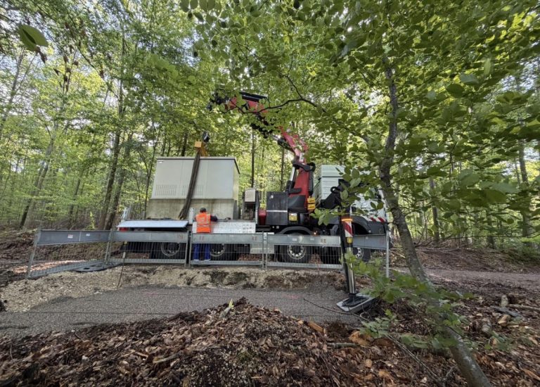 LKW mit Ladekran beim Setzen einer Trafostation im Wald, davor ein vorbereitetes Splittplanum und eine Baustellenabsperrung.