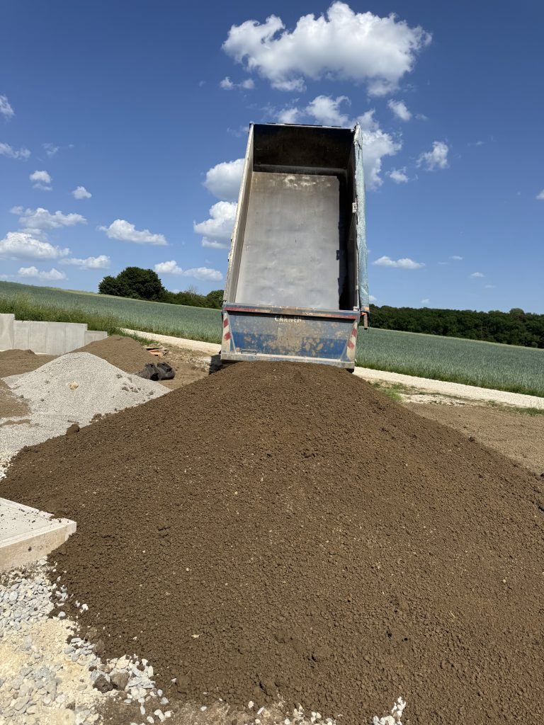 LKW mit hochgestellter Kippmulde hinter einem frisch abgeladenen Erdhaufen auf einer Lagerfläche.