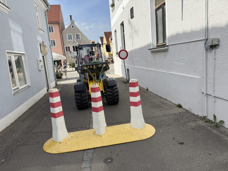 Kompakter Radlader hinter einer mobilen Absperrung mit drei rot-weißen Pfosten in einer schmalen Gasse zwischen Gebäuden.