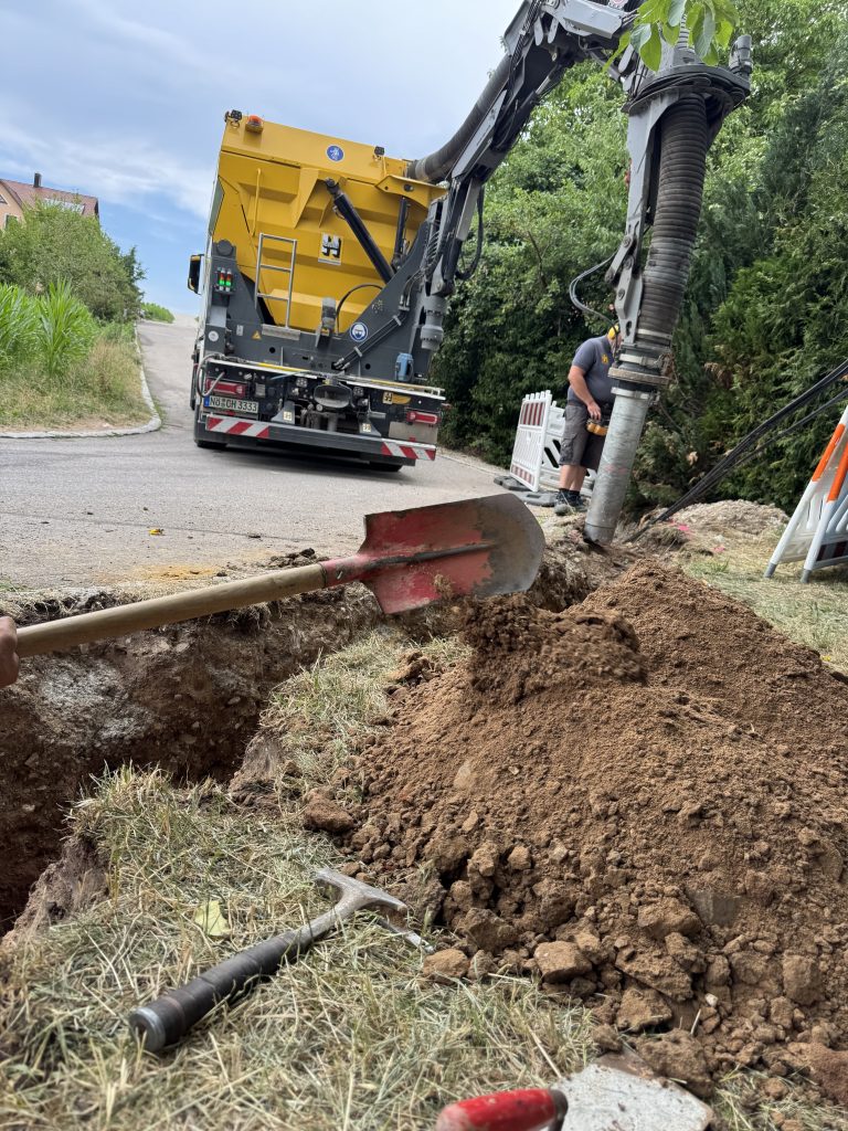 Saugbagger mit abgesenktem Saugschlauch beim Freilegen einer Leitungstrasse am Straßenrand, daneben ein Mitarbeiter.