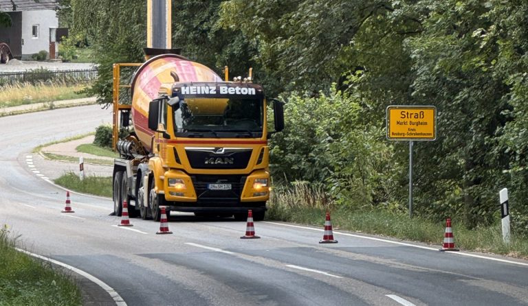 Gelber Betonmischer mit ausgefahrenem Förderband auf einer durch Leitkegel abgegrenzten Fahrbahn am Ortseingang von Straß.