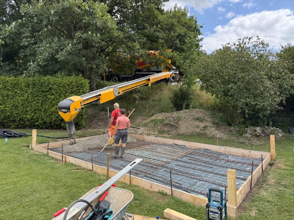Zwei Mitarbeiter beim Betonieren einer Bodenplatte für eine Gartenhütte mit Holzschalung, Bewehrungsmatten und Betonförderband.
