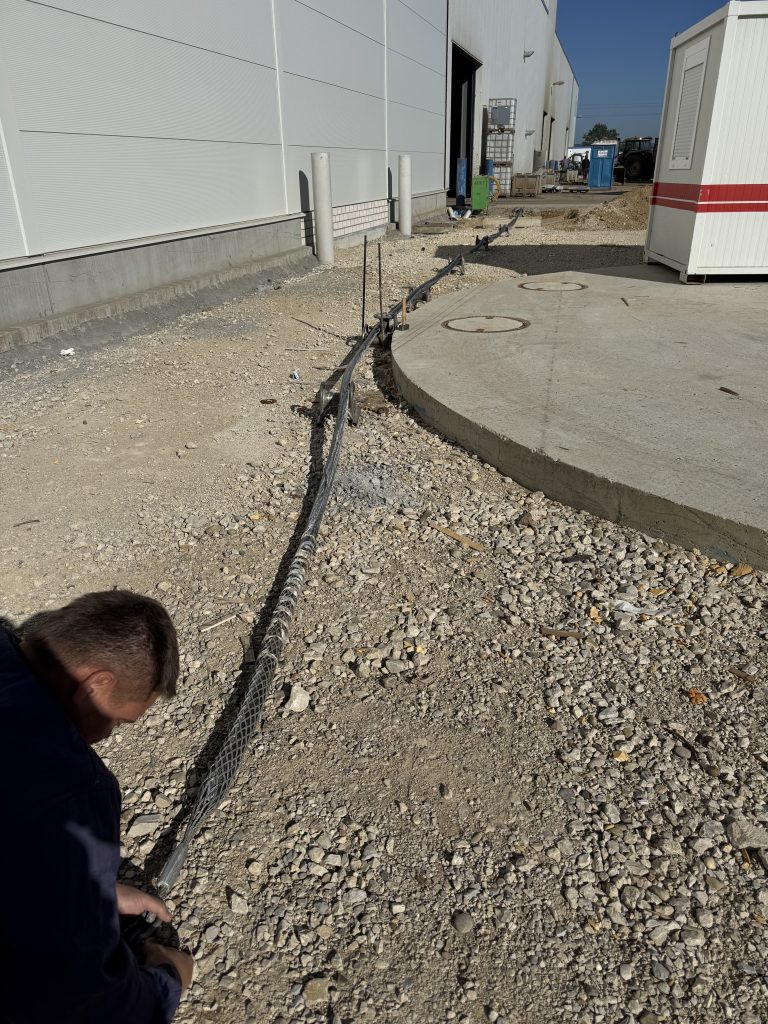 Mitarbeiter am Kabel mit Kabelziehstrumpf auf einer geschotterten Baustellenfläche neben einem Gewerbegebäude.