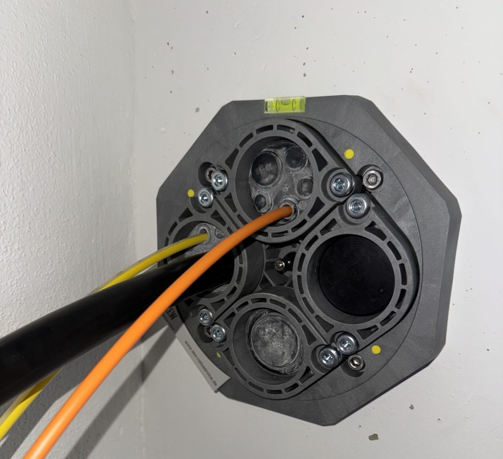 Mehrspartenhauseinführung in einer Kellerwand mit eingeführtem Stromkabel und orangefarbenem Glasfaser-Leerrohr.