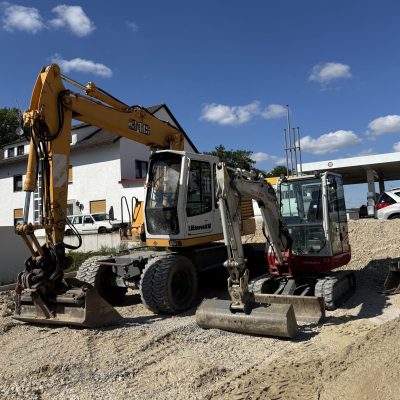 Mobilbagger und Minibagger auf einer geschotterten Baustellenfläche mit Anbaugeräten und Reifenspuren im Vordergrund.